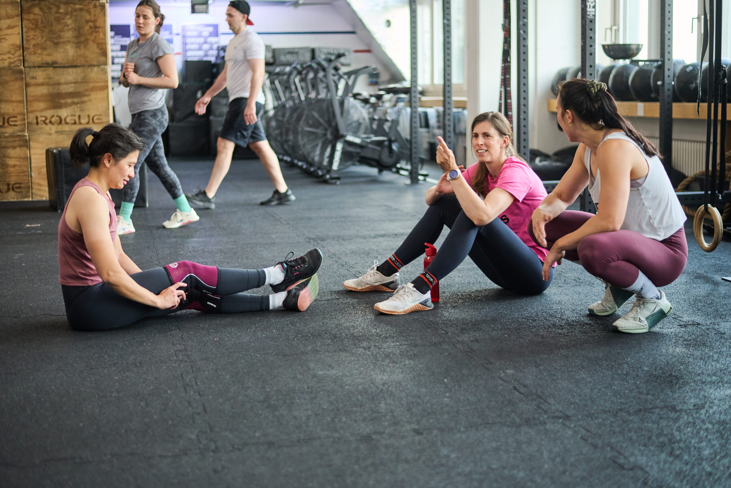 Three women sitting on the gym floor, smiling and recovering after an intense CrossFit workout.